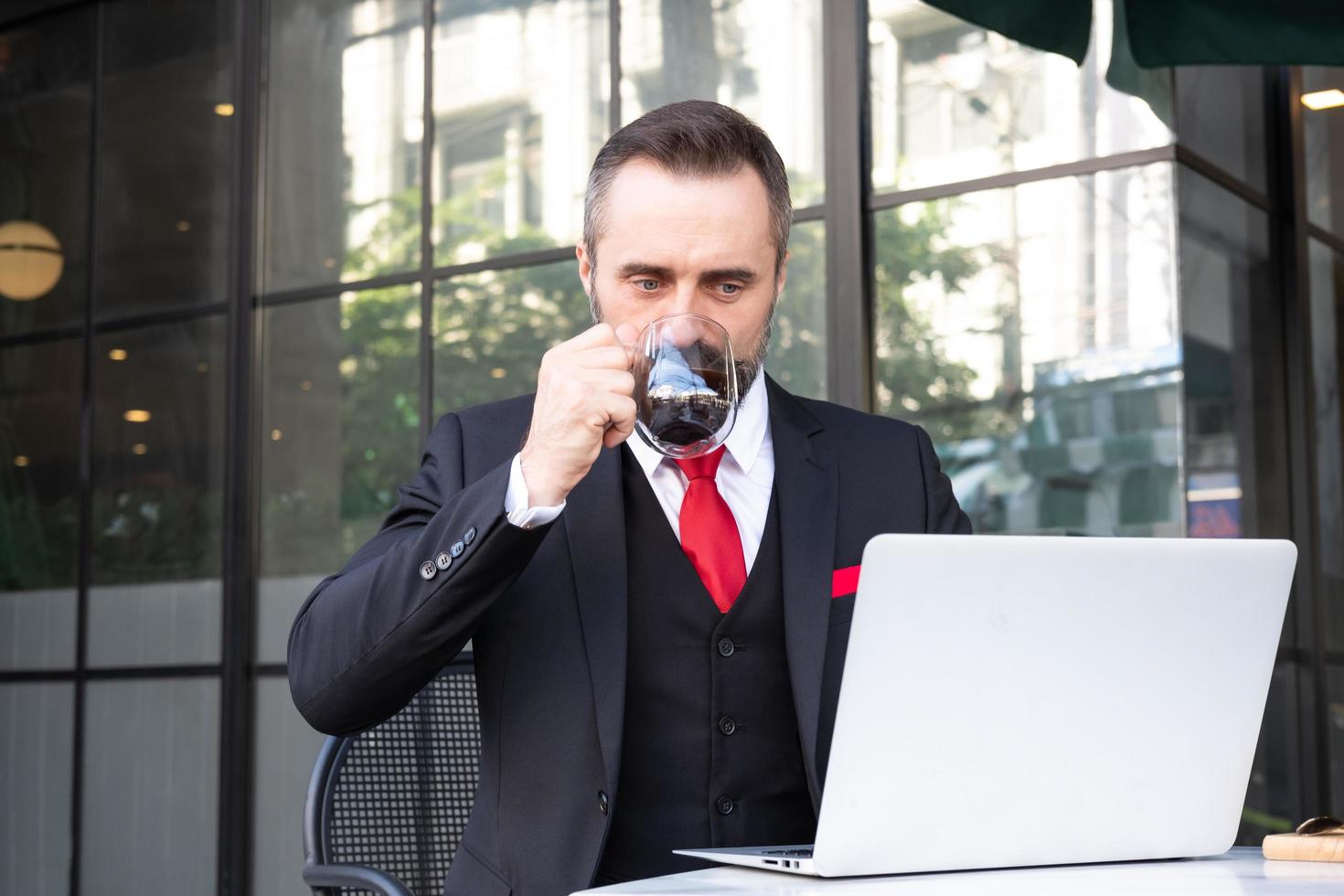 Outdoor shot of Caucasian businessman drinking hot morning coffee at the coffee shop while using computer laptop to work and check his email.  Working routine with urban business area background. photo