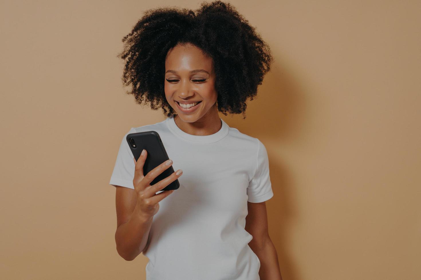 Happy african female student using mobile phone while standing isolated over sand color background photo