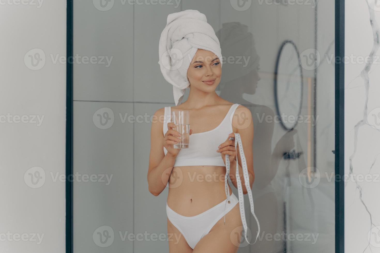 Fit beautiful young girl in bathroom with glass of fresh water and measuring tape photo