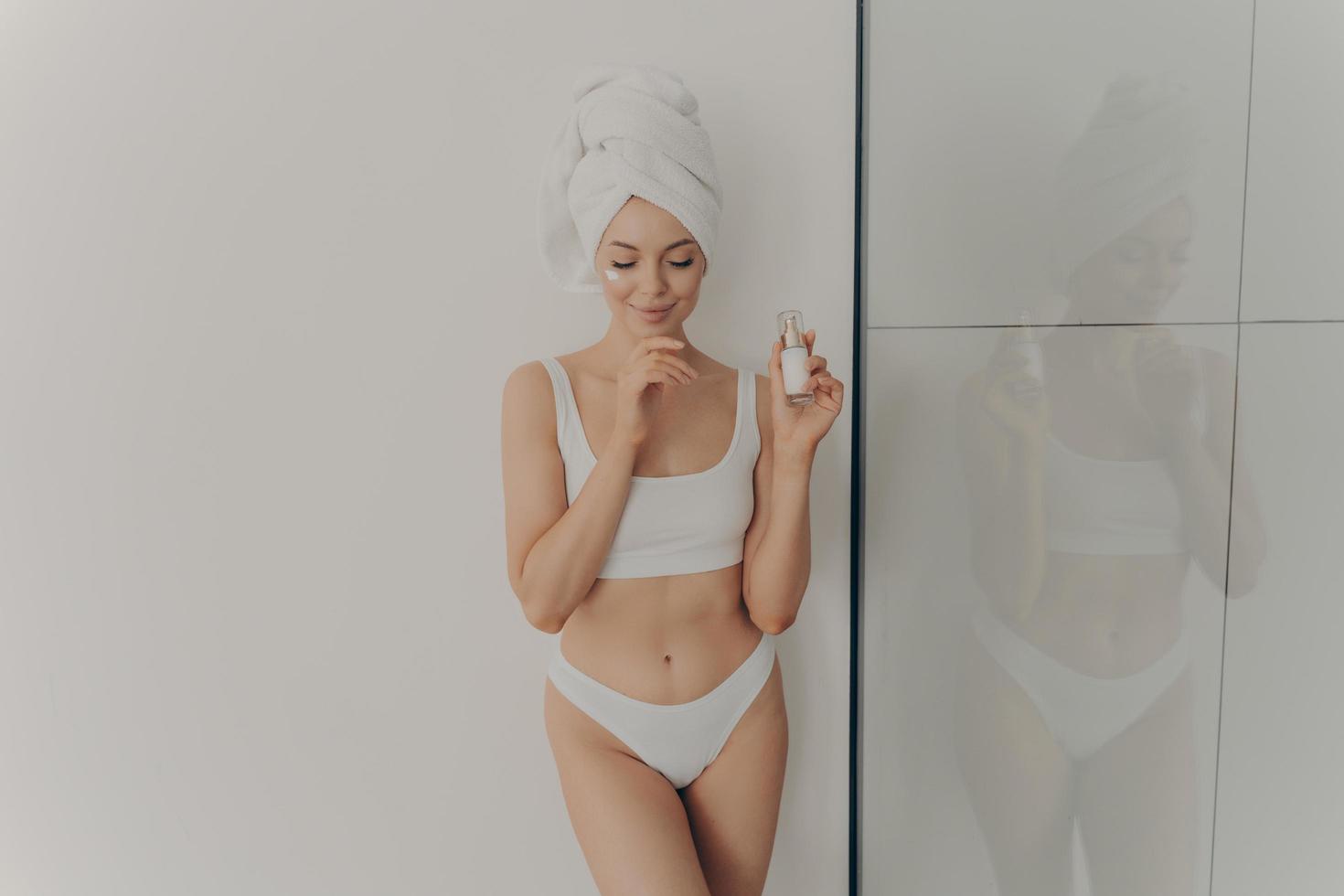 Young girl posing with cosmetic moisturizing face cream or serum in hand isolated over light wall in bathroom photo