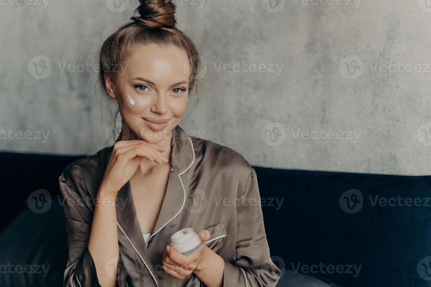 Attractive young female model in brown silk pajama sitting on bed with face cream photo