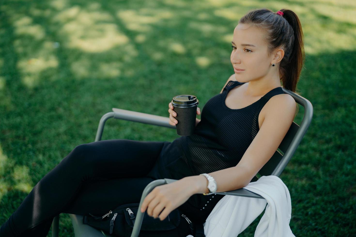 Outdoor shot of thoughtful dark haired beautiful female dressed in sports clothes drinks takeaway coffee spends free time in open air poses on chair at garden enjoys good weather breathes fresh air photo