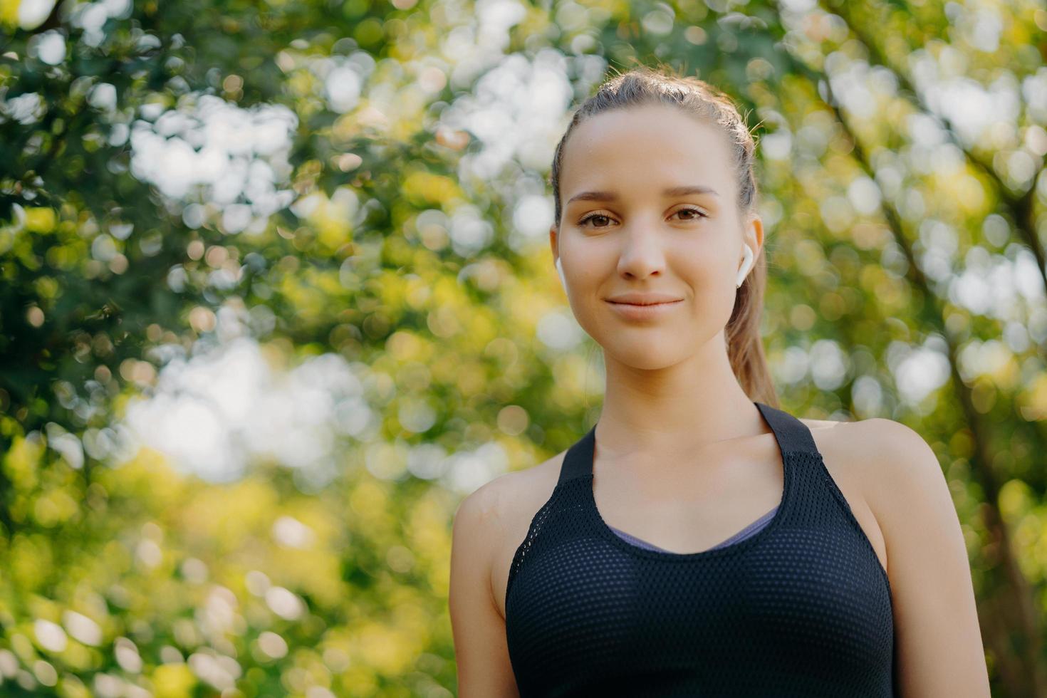 foto de una hermosa mujer deportiva en ropa deportiva usa auriculares inalámbricos para escuchar música descansa en un parque verde mira directamente a la cámara descansa después de un entrenamiento al aire libre involucrado en actividades deportivas