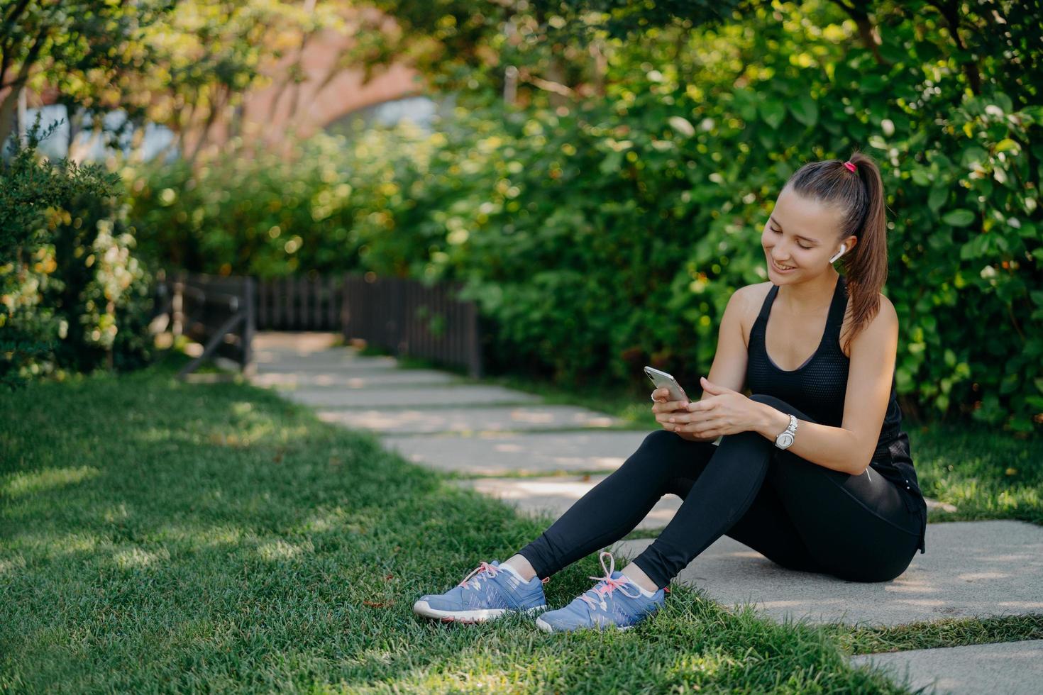 Pleased athletic woman in sportsclothes smiles cheerfully surfs mobile phone while listens music via wireless earphones takes break after sunny morning workout uses app for activity tracking photo