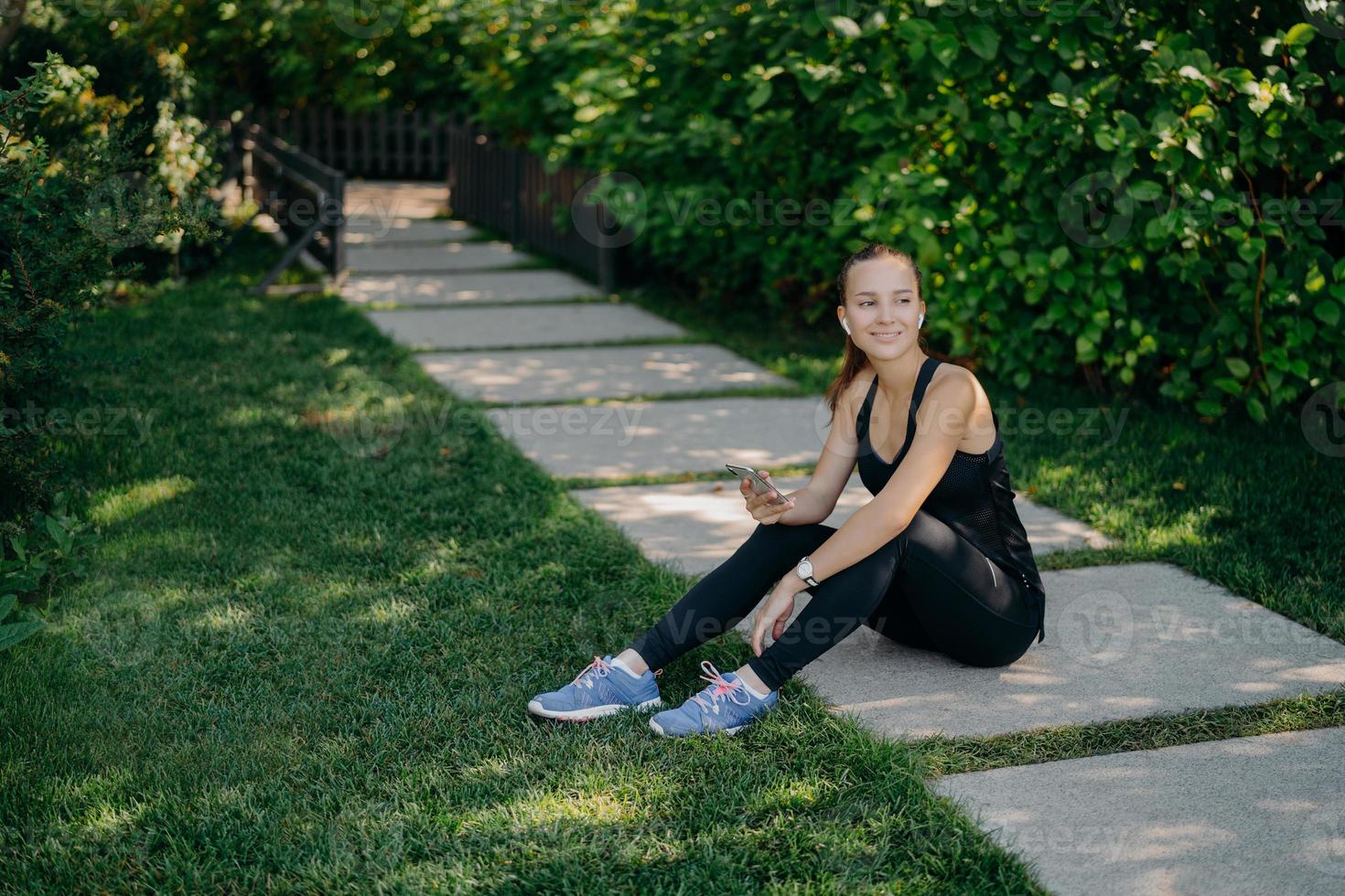 Outdoor shot of active brunette European woman downloads music on smartphone for active training outside has break after workout dressed in active wear shares content in blog looks happily away photo