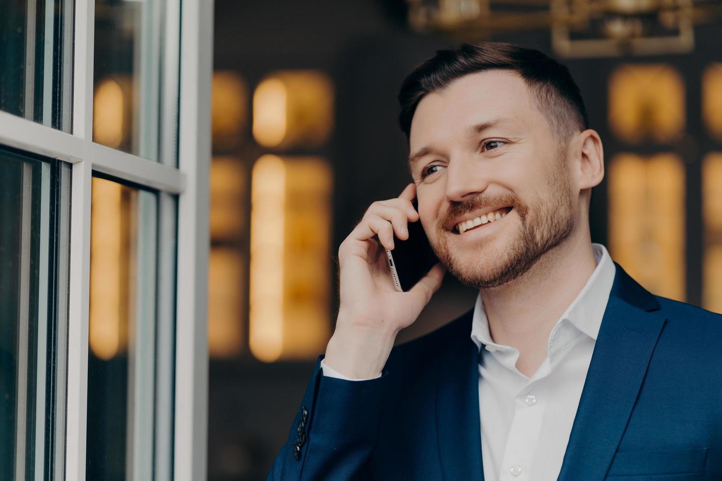 Mobile call. Handsome male executive manager with bristle has telephone conversation enjoys modern communication smiles happily looks thoughtfully into distance wers white shirt and elegant suit photo