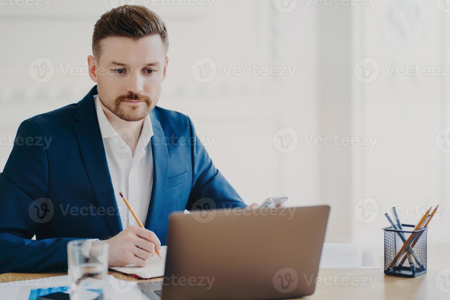 Serious bearded man marketer involved in working process makes notes with pencil looks attentively at laptop computer wears blue formal suit poses at coworking space writes organisation plan photo