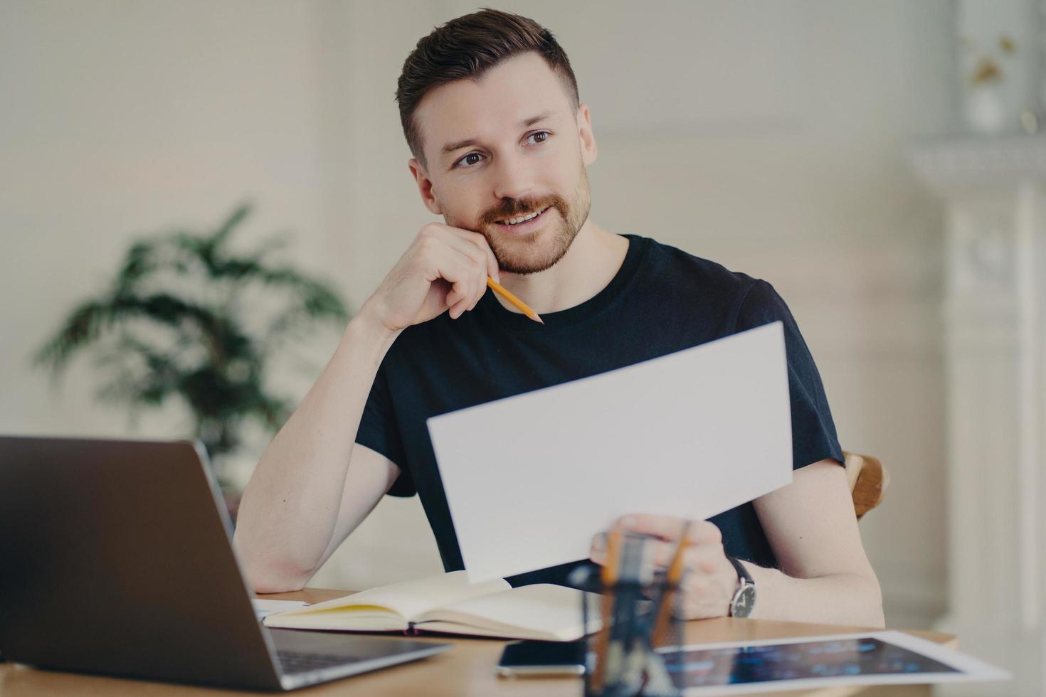 Glad handsome European businessman holds paper document poses in home interior at desktop surrounded by modern technologies checks information has remote work looks thoughtfully into distance photo