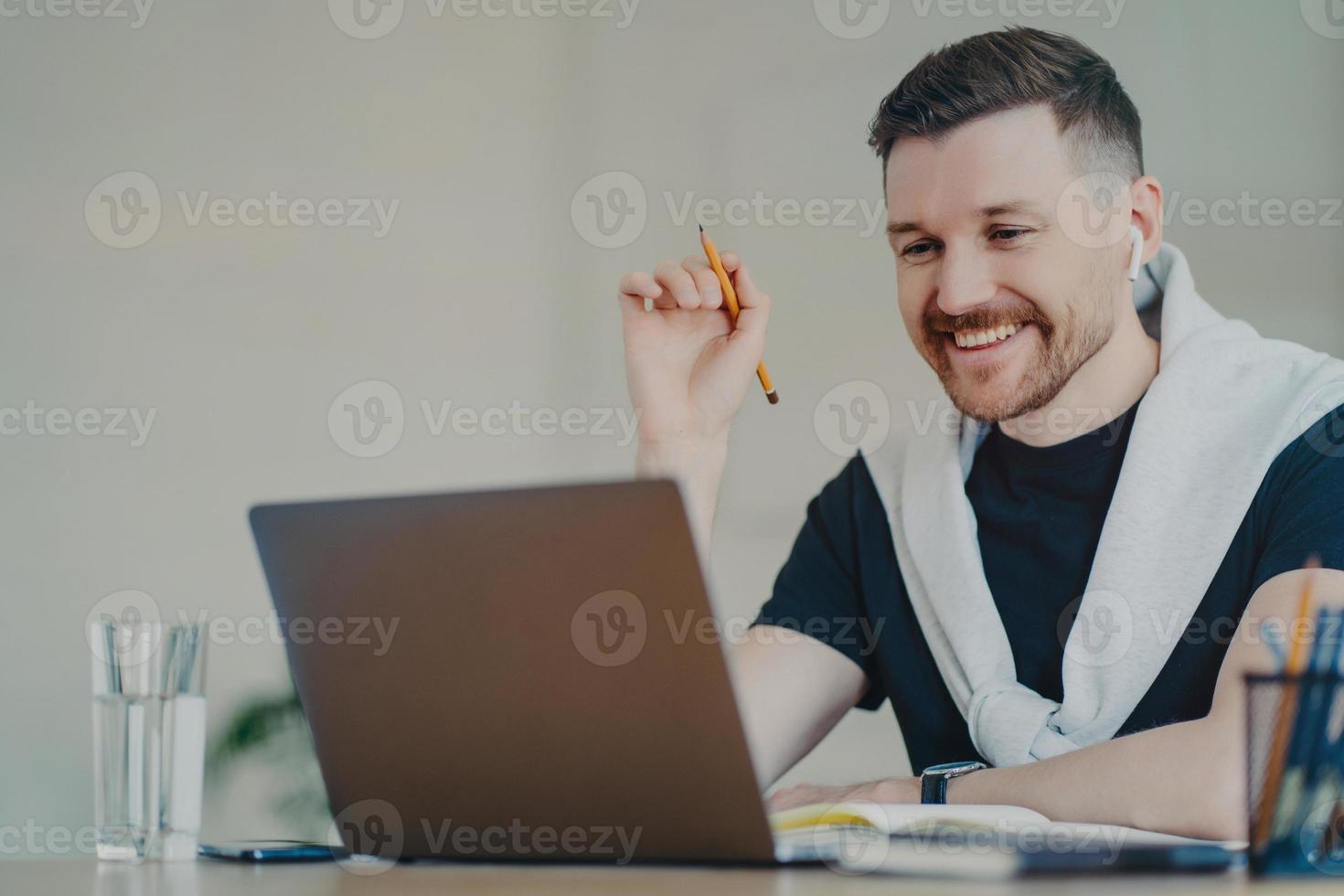 el hombre barbudo feliz prepara el trabajo del curso en papel trabaja de forma remota desde casa ve el seminario web utiliza una conexión a Internet de alta velocidad una computadora portátil moderna y los auriculares hablan con un compañero en línea. tiempo de trabajo. foto