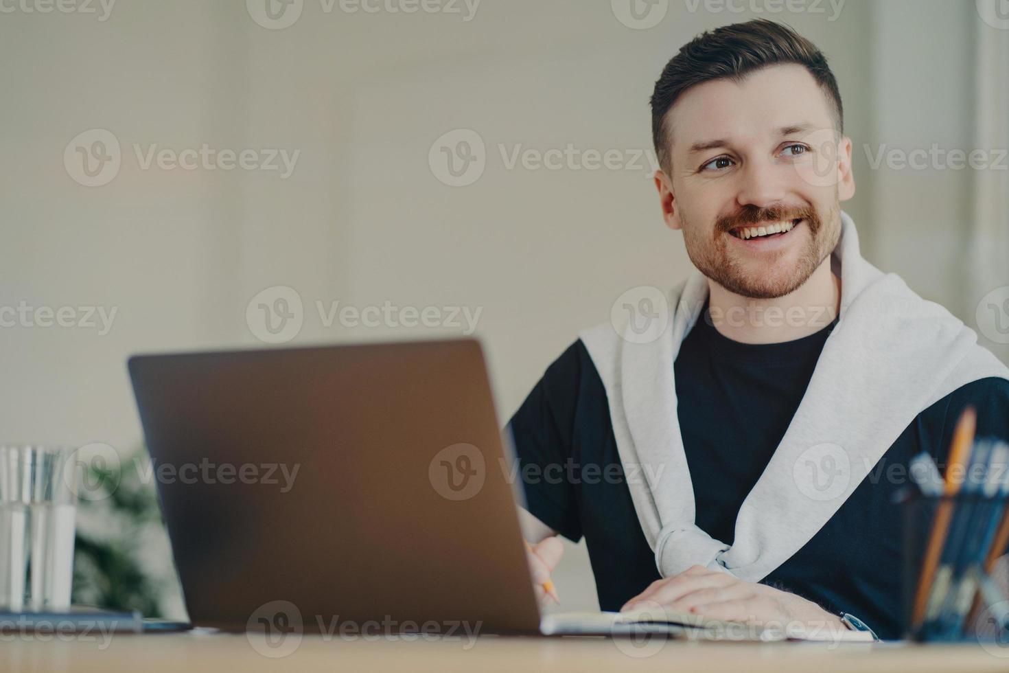 Happy bearded man office worker writes organisation plan works remotely makes notes of info sits in front of opened laptop looks away dressed in casual t shirt and jumper tied over shoulders photo
