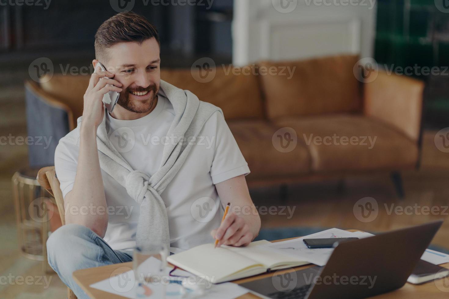 Cheerful man freelancer makes telephone call writes down information in notepad makes notes in planner looks attentively at laptop screen poses against home interior at desktop. Technology user photo