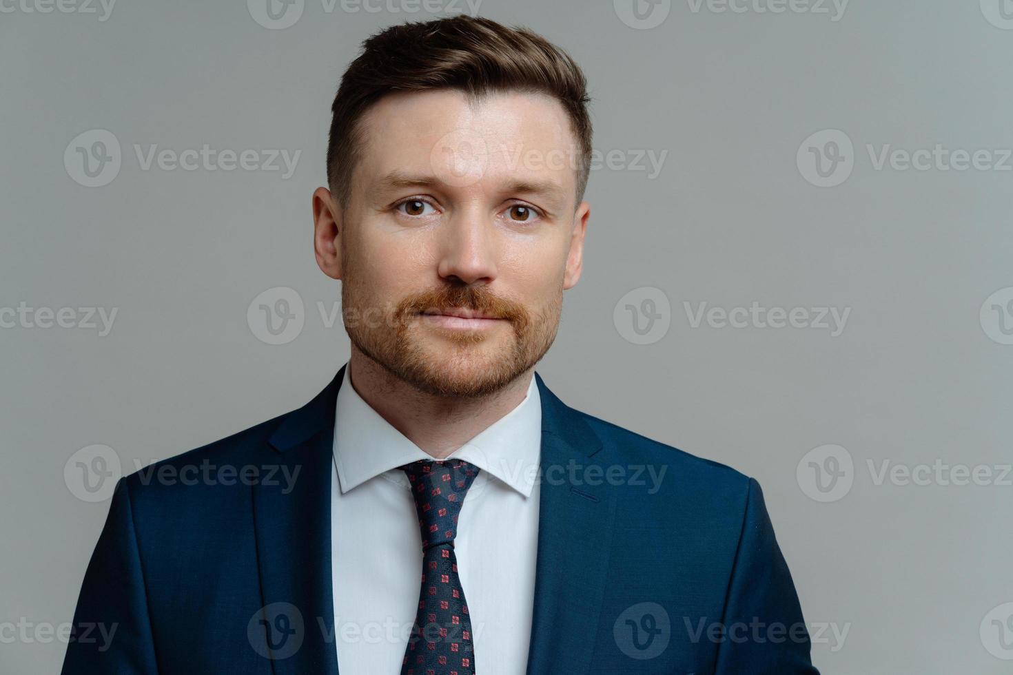 Serious confident businessman wears formal clothes looks directly at camera has well groomed appearance ready for meeting with partners isolated over grey studio background. Motivated entrepreneur photo