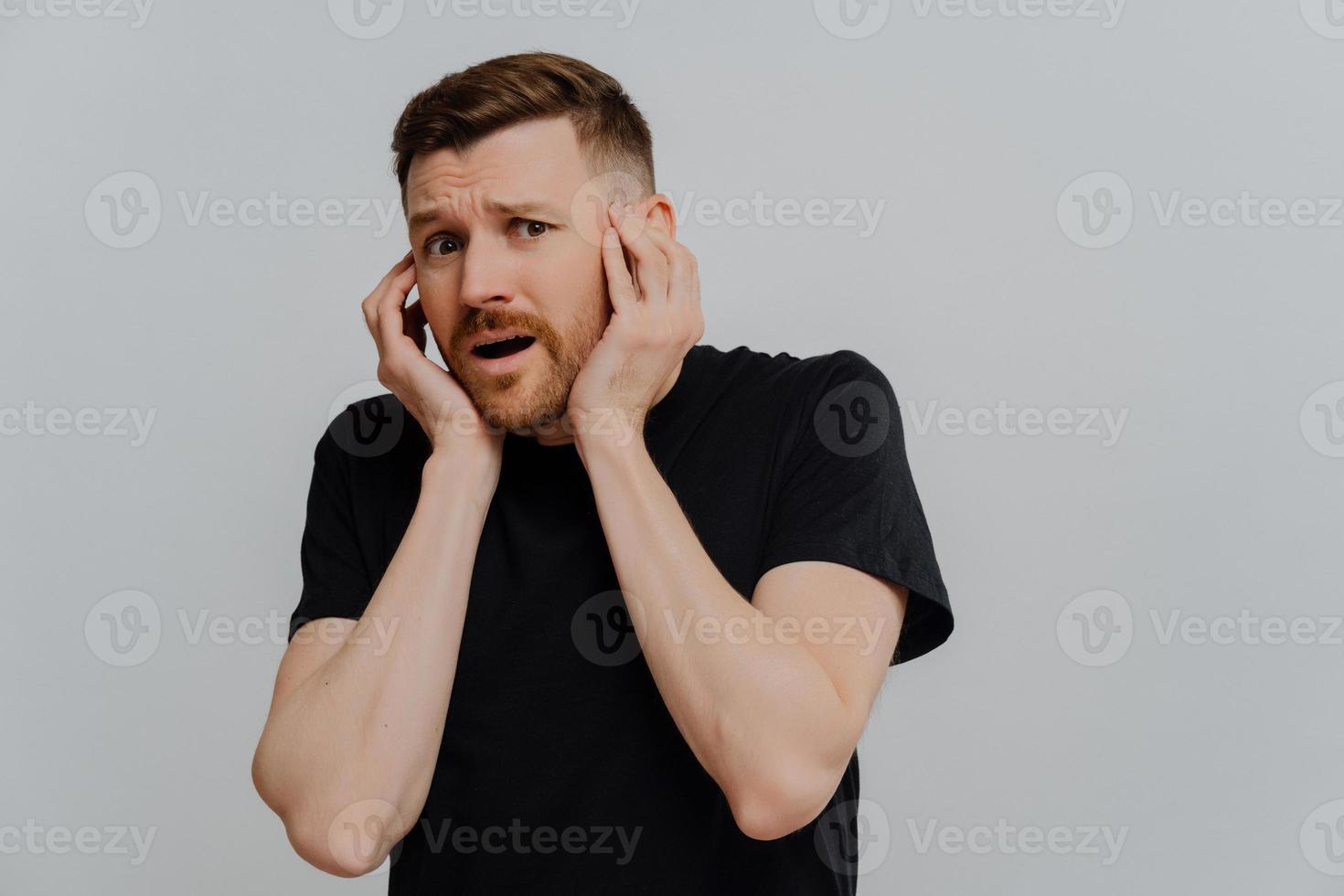 Scared frightened young European man keeps hands on face being afraids of horrible scene wears casual black t shirt gasps from fright isolated over blue studio background. Omg how scary it is. photo