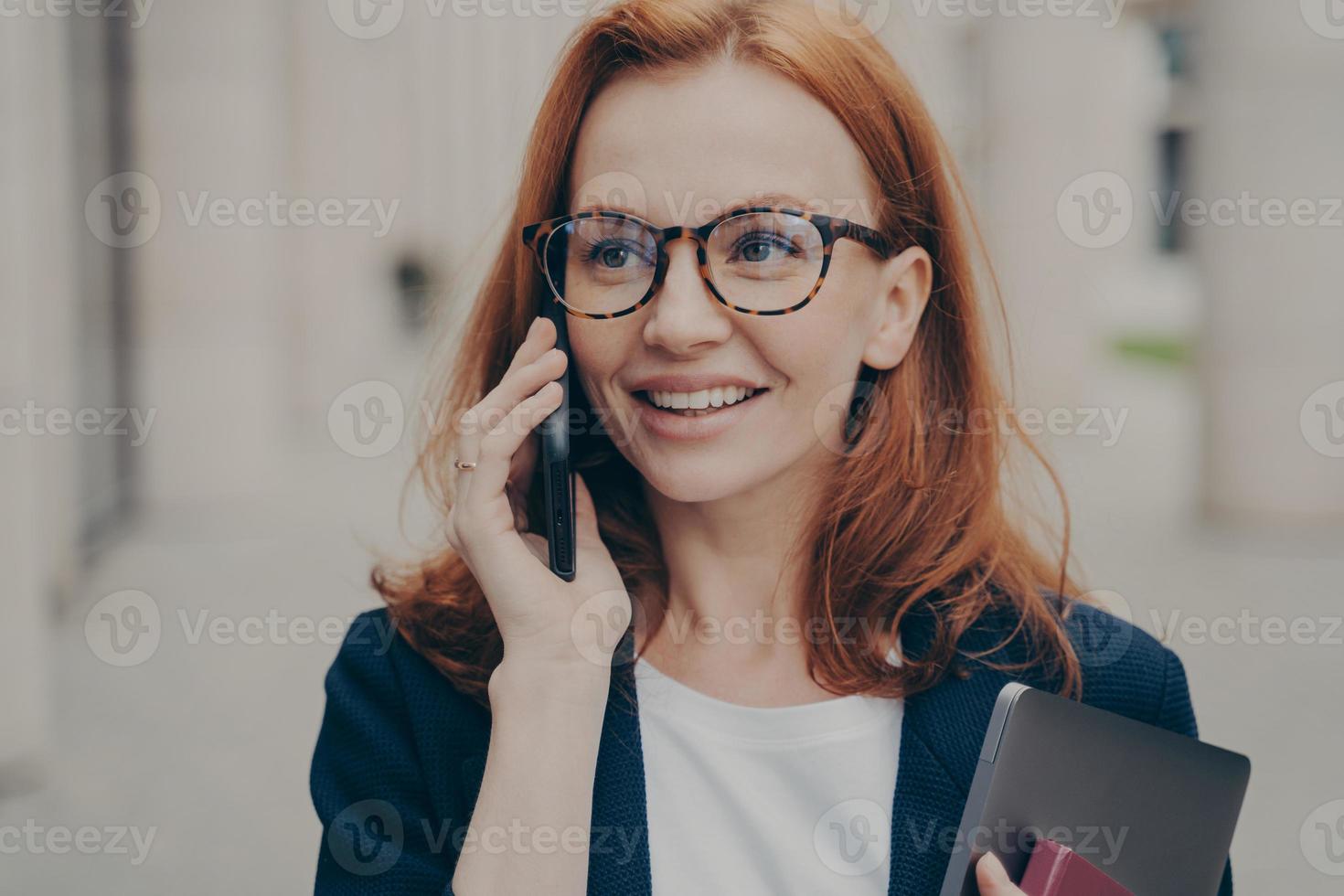 sonriente mujer de negocios pelirroja positiva con gafas llamando a su pareja, de pie al aire libre foto