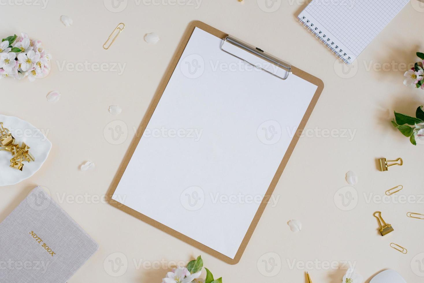 hoja de papel en blanco con un clip, ramas de manzano con flores, blocs de notas y clips de papel sobre un fondo beige. un escritorio mínimo de oficina en casa. endecha plana, vista superior. foto