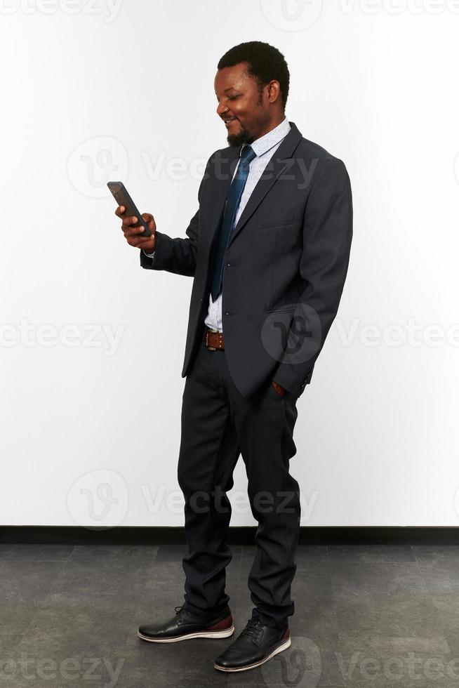 Smiling african american black man in business suit looks in smartphone white wall background photo