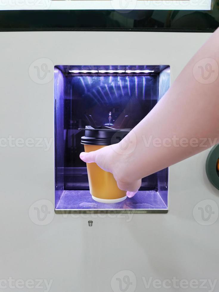 One hand is holding a yellow paper cup at the window of a coin-operated coffee machine. photo