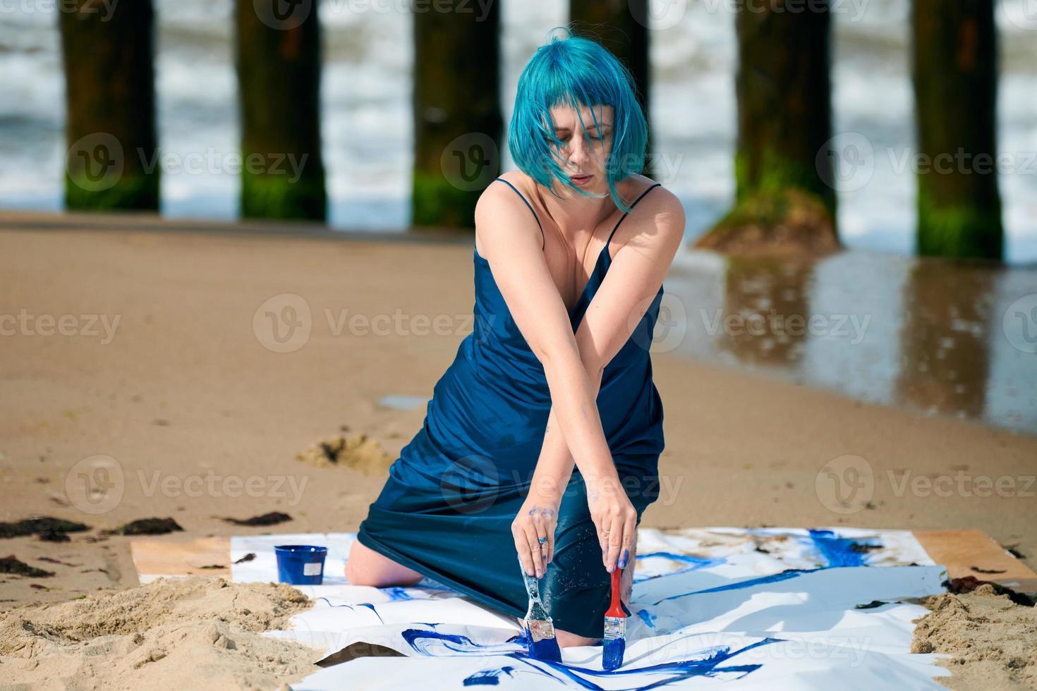 Artistic blue-haired woman performance artist smeared with gouache paints on large canvas on beach photo