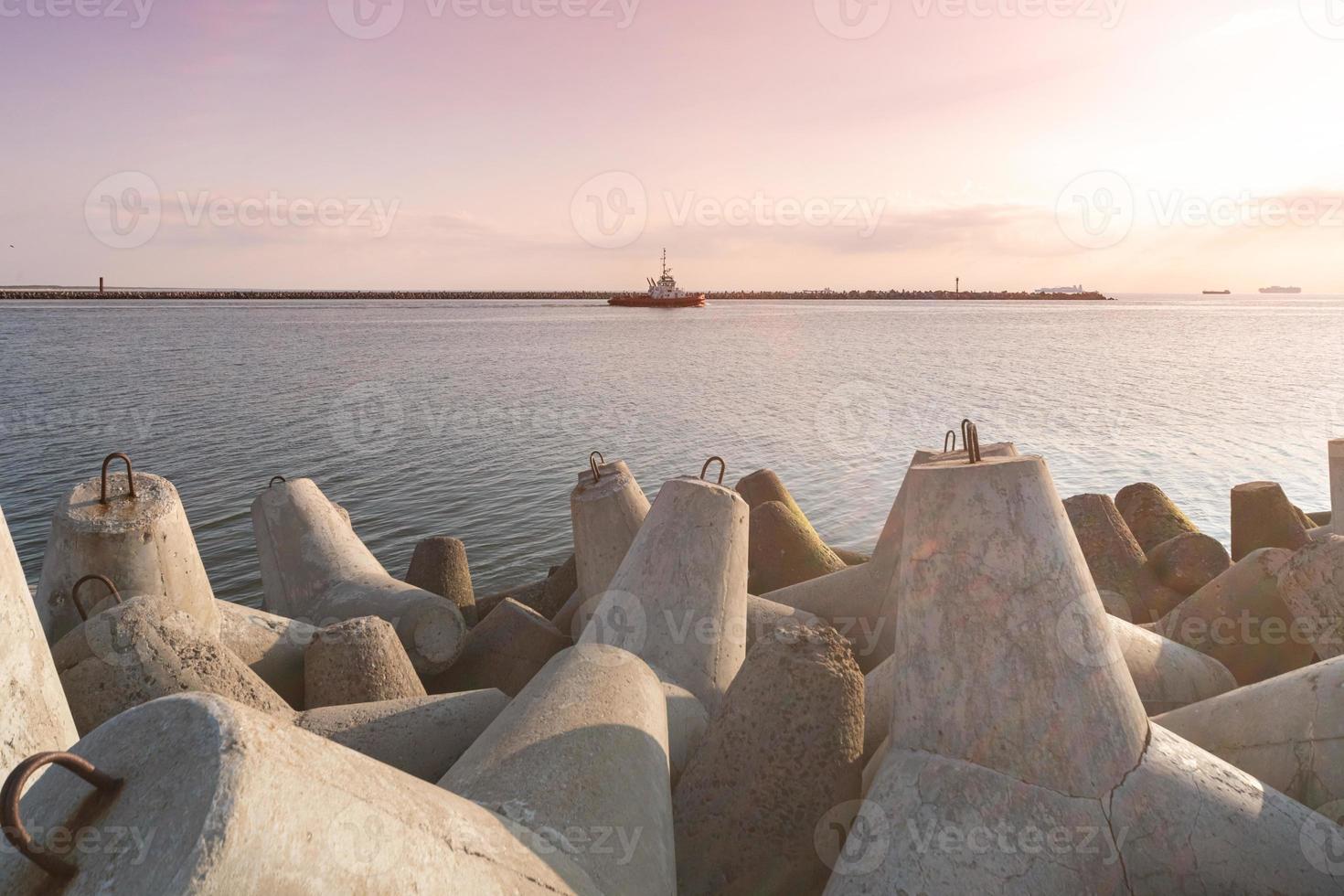 Tugboat sailing in sea to tow ship to port. Tetrapod breakwaters in harbor. Beautiful sunset over the pier. photo