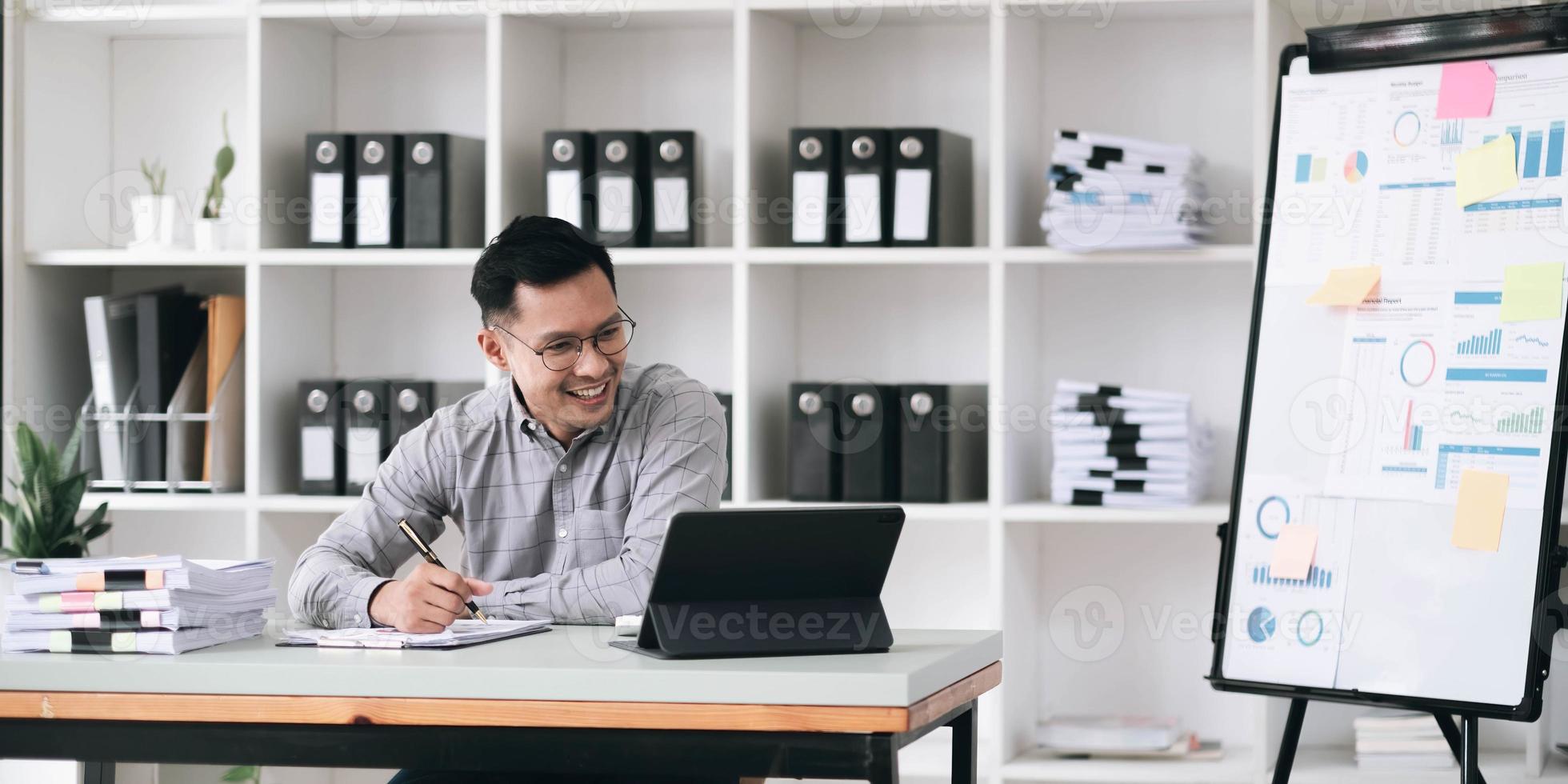 Portrait of a young Asian businessman smiling while using a laptop and writing down notes while sitting at his desk in a modern office photo