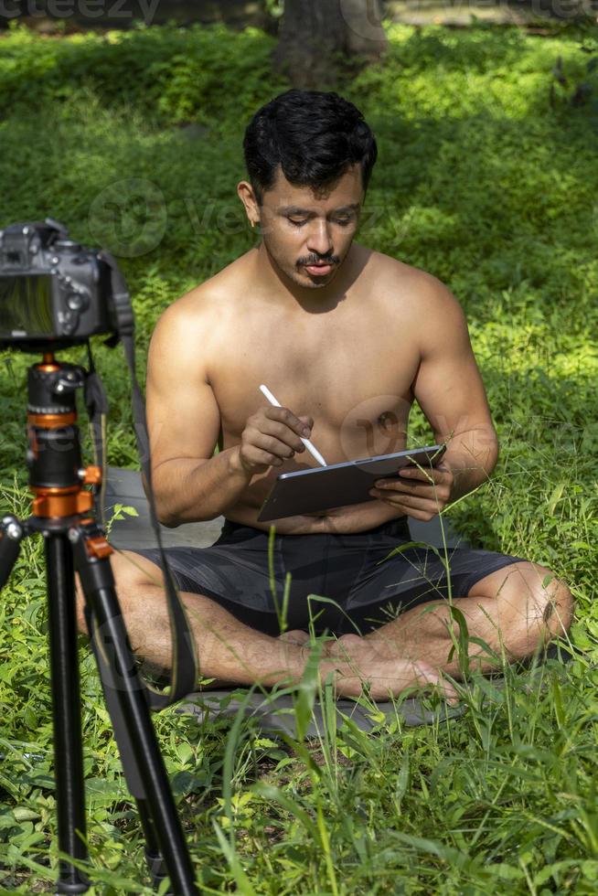Young man practicing yoga asana, balance, meditating while standing on one leg on sports mat on green grass in park. Using tablet for online class. photo