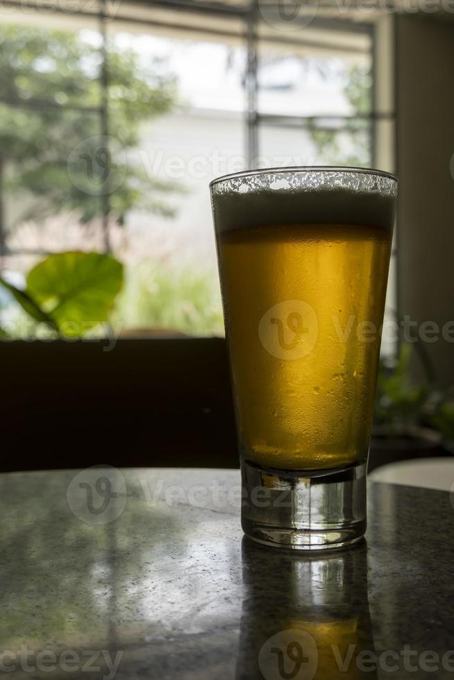 vaso de cerveza en una mesa de terrazo, dos sillas amarillas en el fondo, mesa retro, ventana con luz natural foto