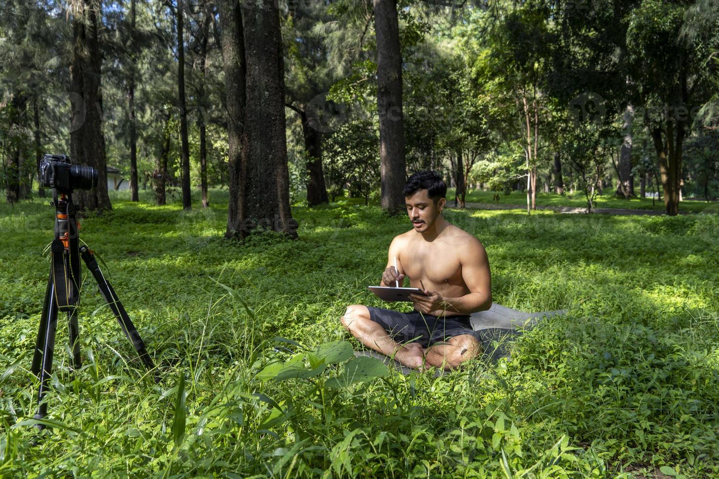 Young man practicing yoga asana, balance, meditating while standing on one leg on sports mat on green grass in park. Using tablet for online class. photo