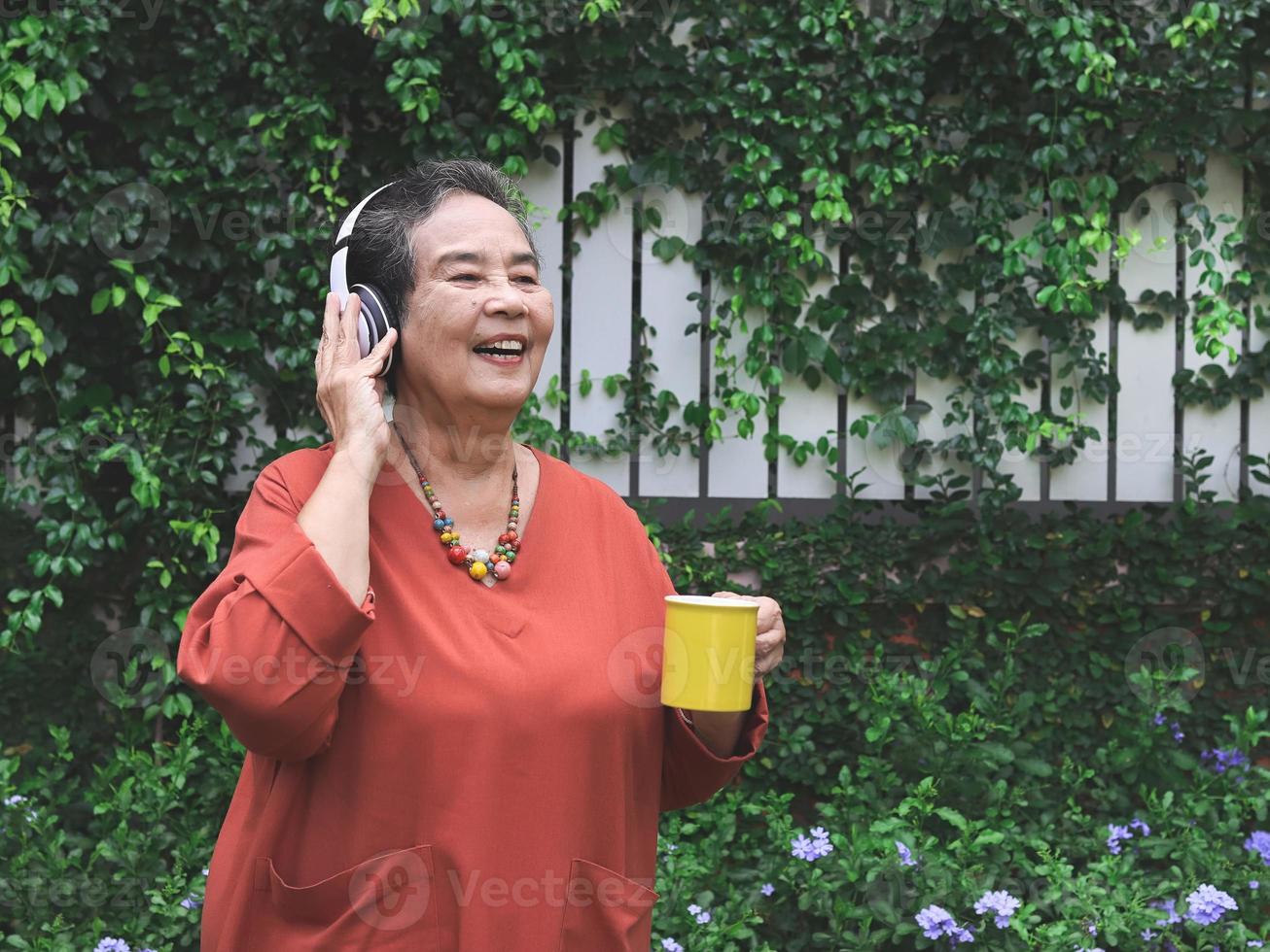 anciana asiática feliz y saludable sosteniendo una taza amarilla de café parada en el jardín, escuchando la música de los auriculares, sonriendo y mirando hacia otro lado. foto