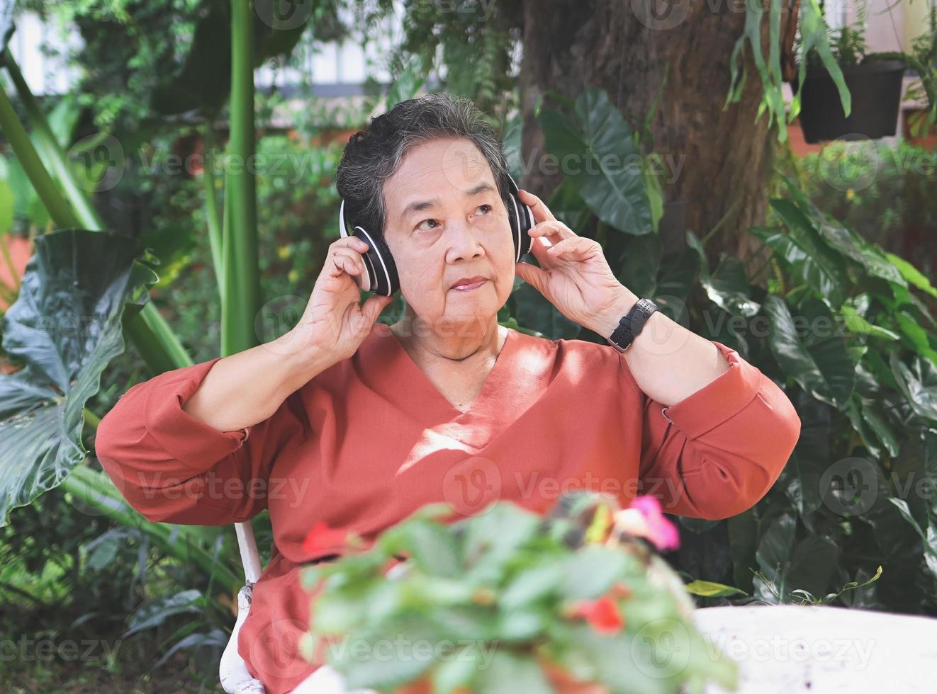 anciana asiática feliz y saludable sentada con una taza de café y macetas de flores en el jardín al aire libre, escuchando su música favorita desde los auriculares, sonriendo y mirando hacia otro lado. foto