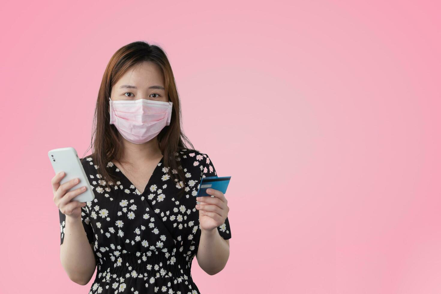 feliz retrato asiático linda mujer joven con máscara quirúrgica sostiene teléfono móvil y tarjeta de crédito de débito de plástico, toma de estudio aislada en fondo rosa, mujer usando compras en línea de teléfonos inteligentes foto