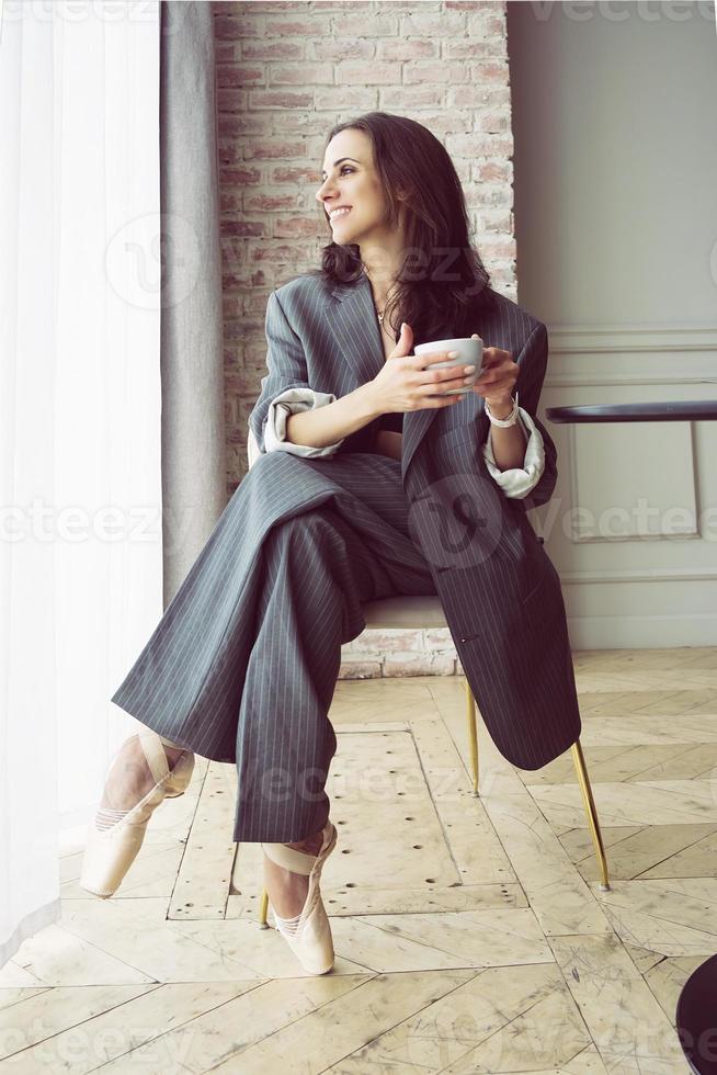 bailarina con traje de rayas grises con una taza de café sonriendo y soñando sentada en una mesa junto a la ventana foto