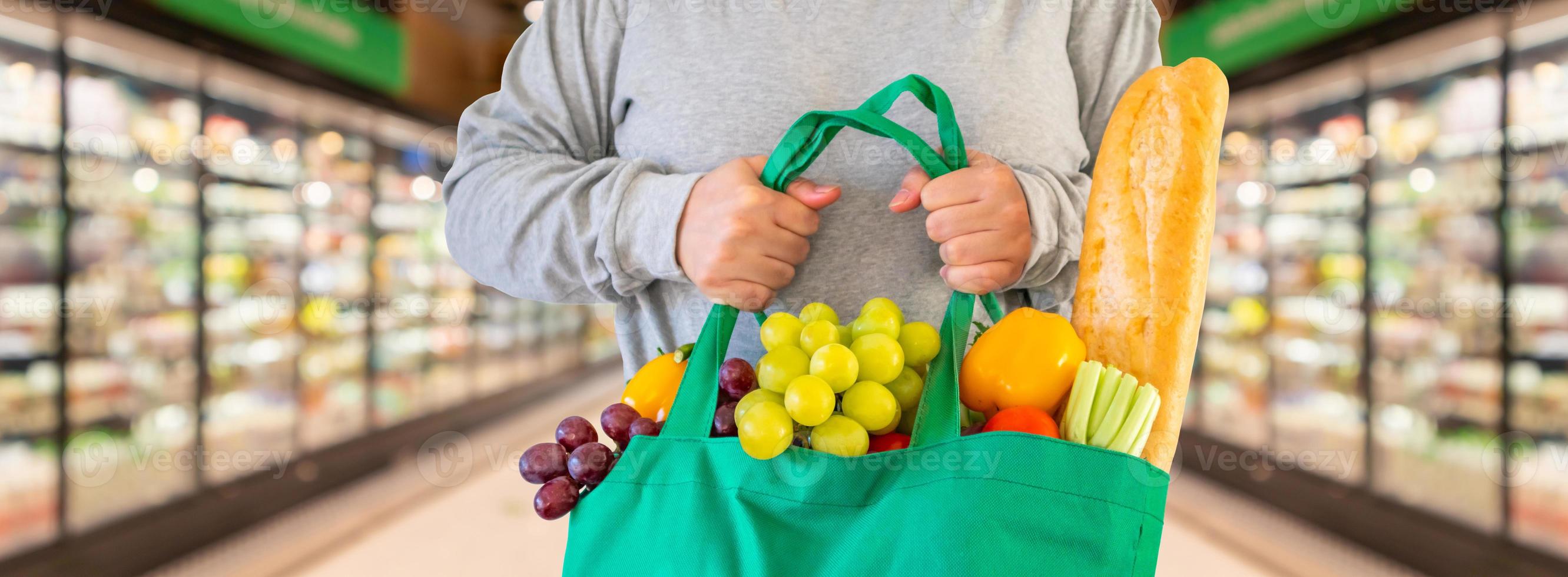 Customer hold reusable green shopping bag with fruit and vegetable with Supermarket aisle background photo