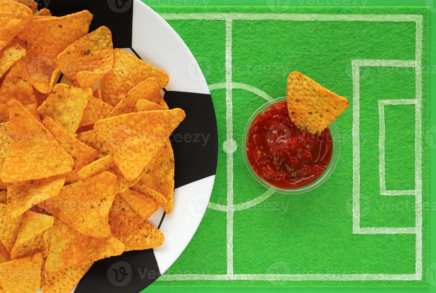 Football soccer party table. Corn nacho chips on big plate painted like soccer ball and one in red salsa sauce on dish mat like football field made of green felt, top view. Watch match broadcast. photo