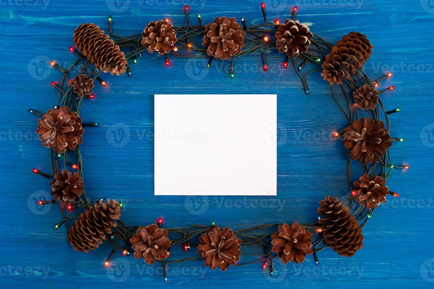 Top view on frame from Christmas lights, pine cones and white sheet of paper on the blue wooden background with copy space. photo