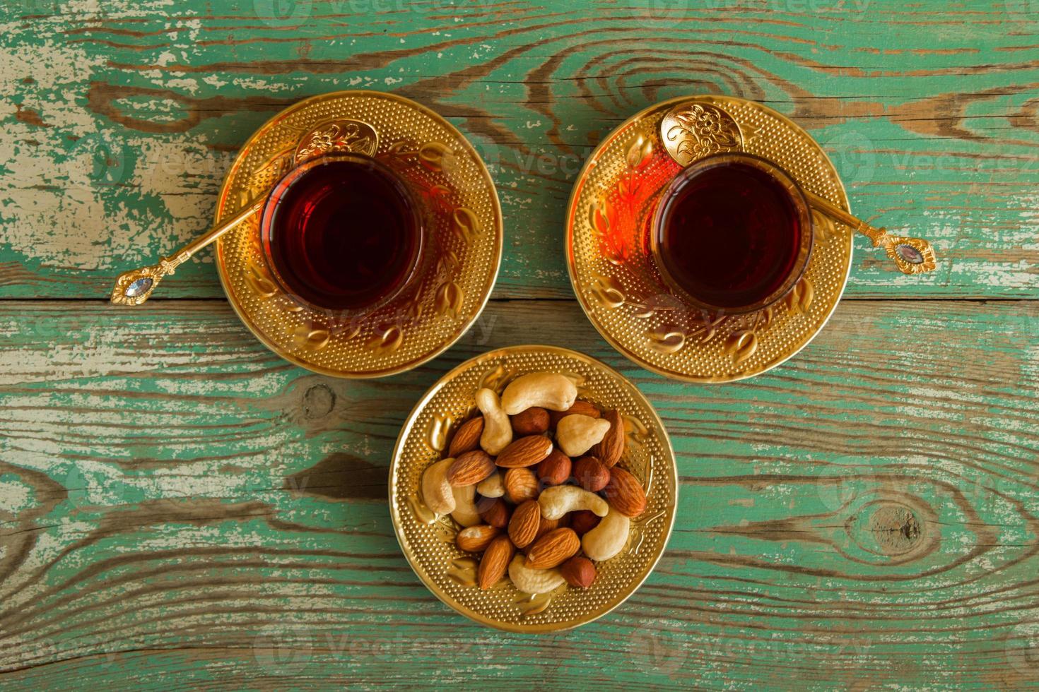 Mixed nuts on a yellow metal saucer and two glass cups of Turkish tea on a turquoise wooden background, top view. photo