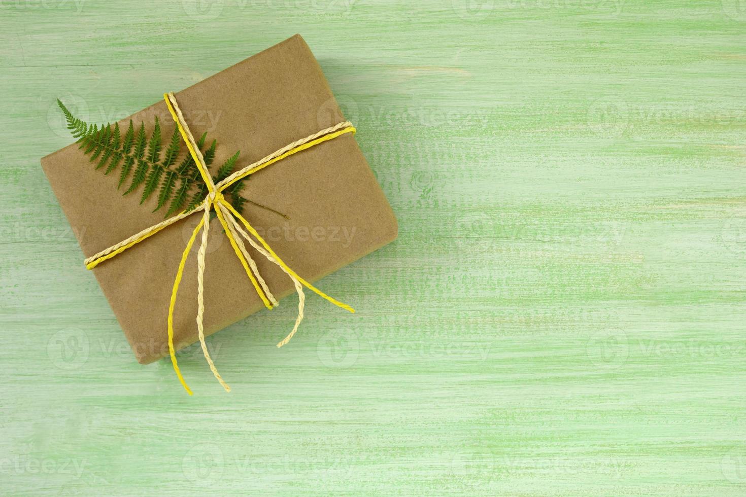 caja de regalo envuelta en papel artesanal y cinta blanca y amarilla con hoja de helecho sobre un fondo de madera verde, vista superior. foto