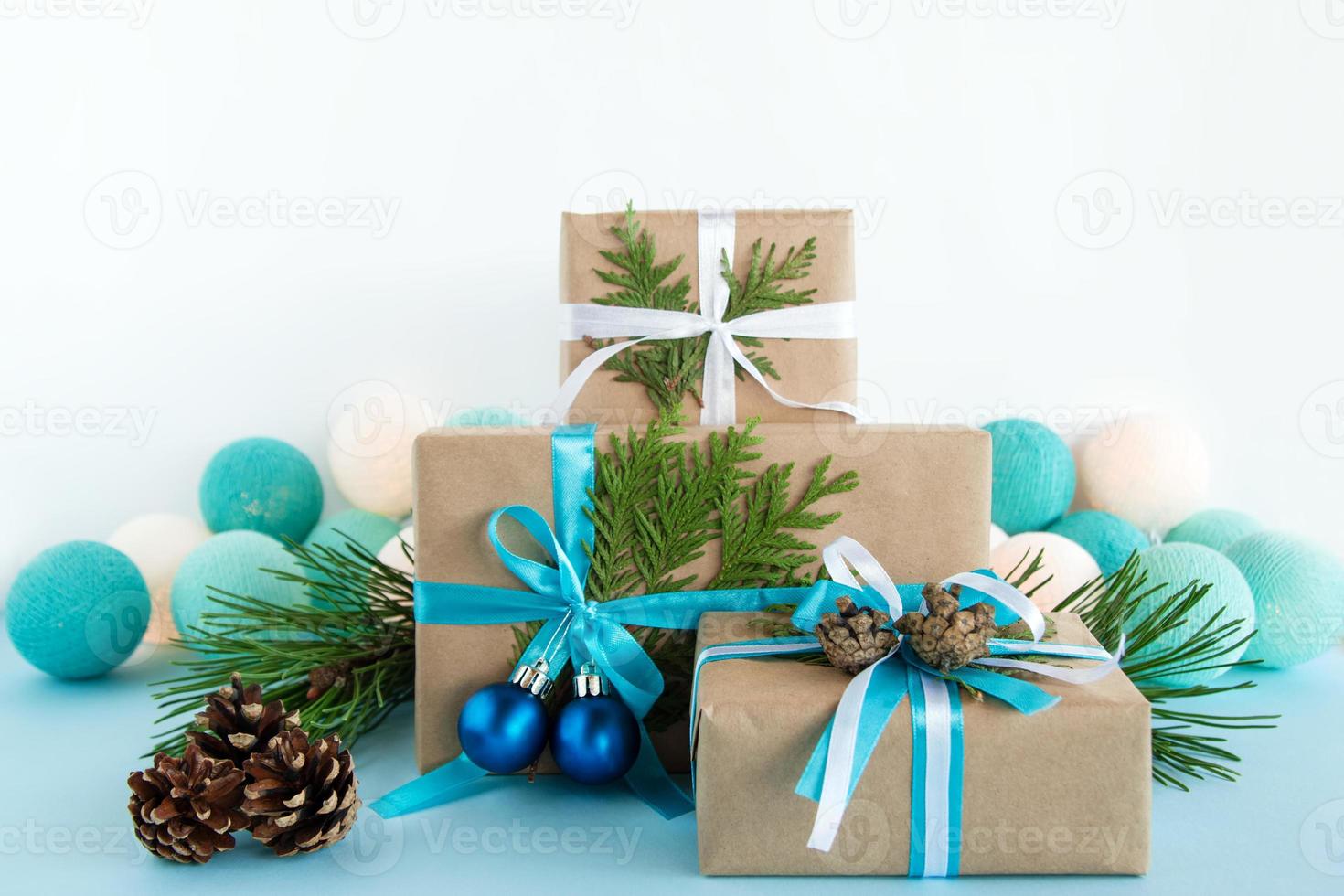 tres cajas de regalo de navidad envueltas en papel artesanal, cintas azules y blancas, decoradas con ramas de abeto, conos de pino y bolas de navidad en el fondo azul y blanco con luces de navidad. foto