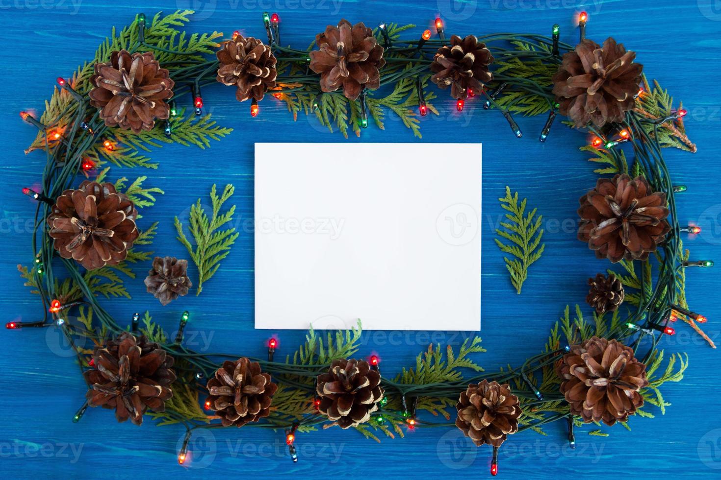 Top view on frame from Christmas lights, fir branches, pine cones and white sheet of paper on the blue wooden background with copy space. photo