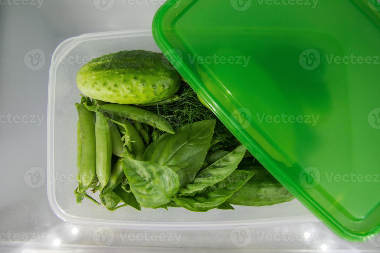 Green vegetable - basil, dill, sorrel, peas, cucumber - in the plastic food container on a shelf of a fridge. photo
