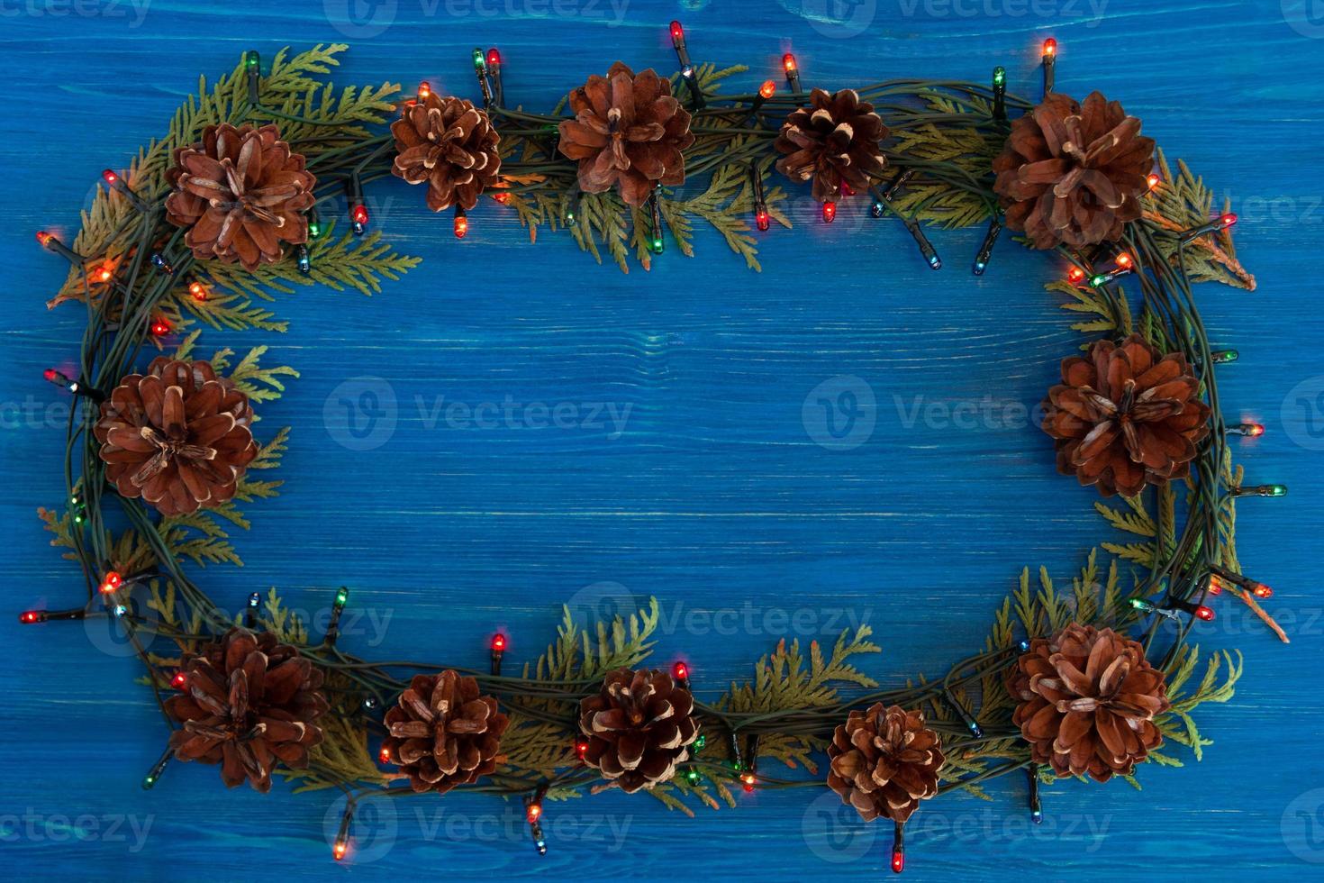 Top view on frame from Christmas lights, fir branches and pine cones on the blue wooden background with copy space. photo