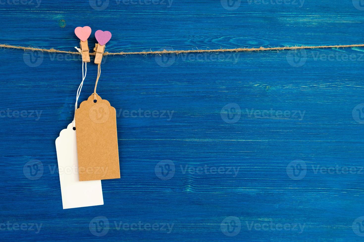Two brown and white blank paper price tags or labels set and wooden pins decorated on hearts hanging on a rope on the blue wooden background. photo