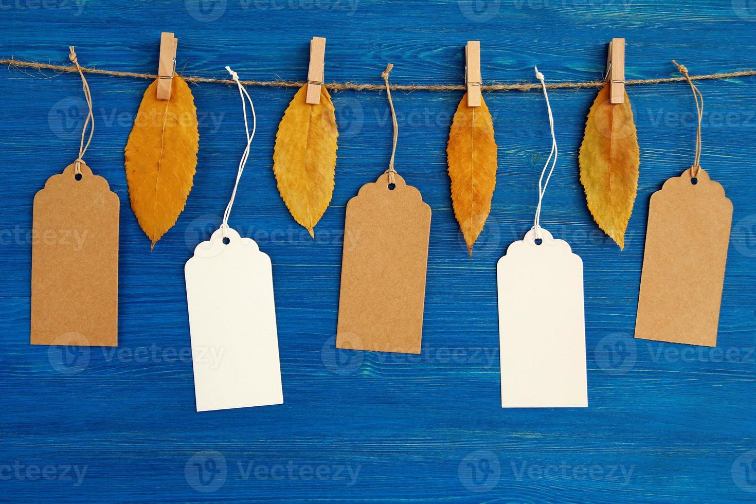 cinco etiquetas o etiquetas de precio de papel en blanco marrón y blanco colgadas en una cuerda con hojas secas de otoño amarillas sobre el fondo de madera azul. foto