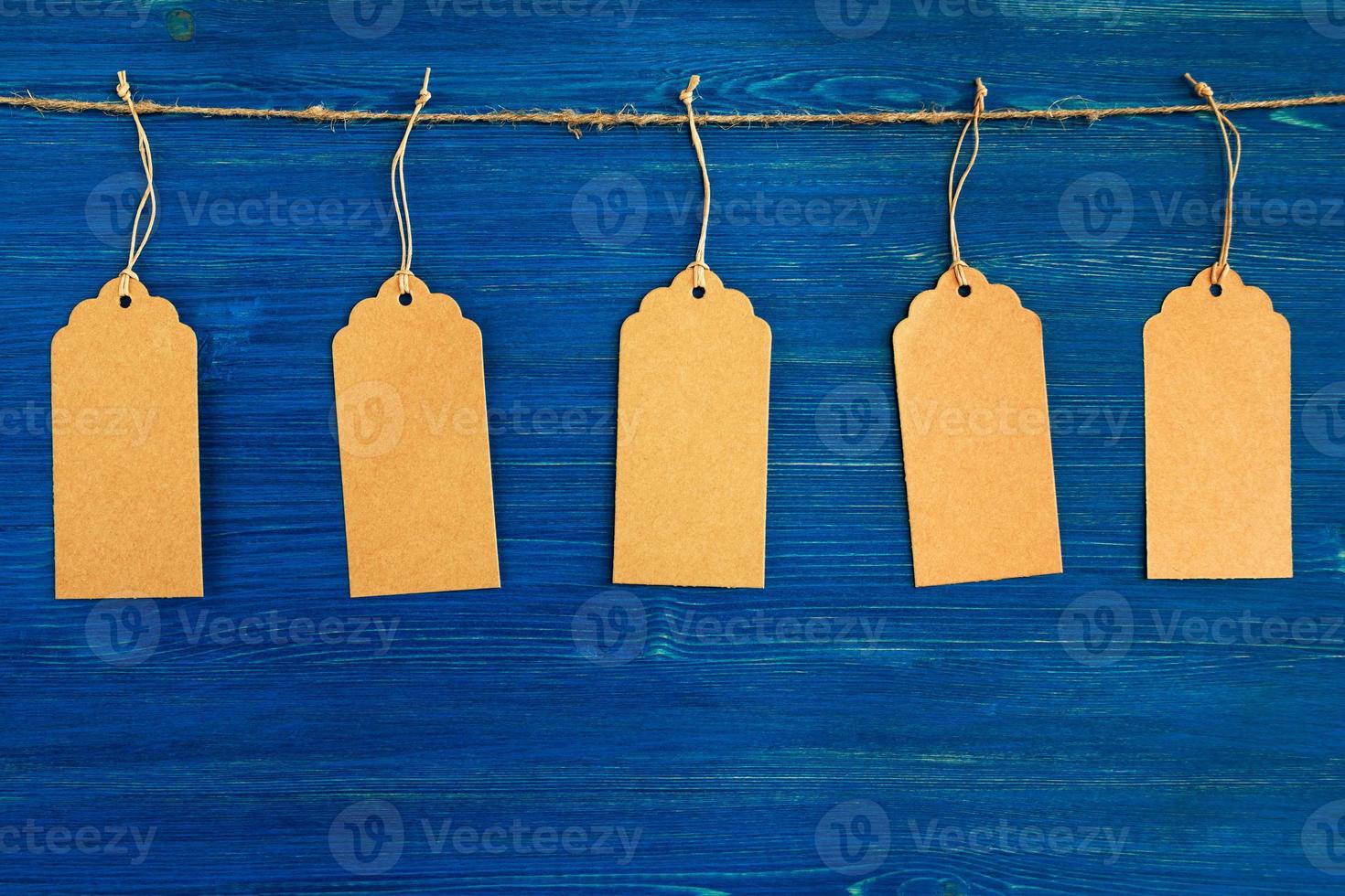 Five brown blank paper price tags or labels set hanging on a rope on the blue wooden background. photo