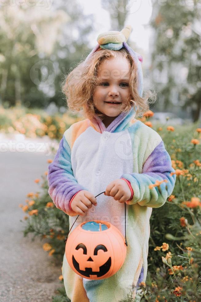 cute little girl in a rainbow unicorn costume for Halloween goes to collect sweets in a pumpkin basket in a residential area. child walks in the outdoor. trick or treat. lifestyle. kigurumi photo