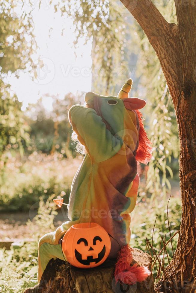 little girl in a rainbow unicorn Halloween costume and a dachshund in a dress with a pumpkin basket for sweets are sitting on stump at forest sunset. fabulous wonderful magical forest. space for text photo