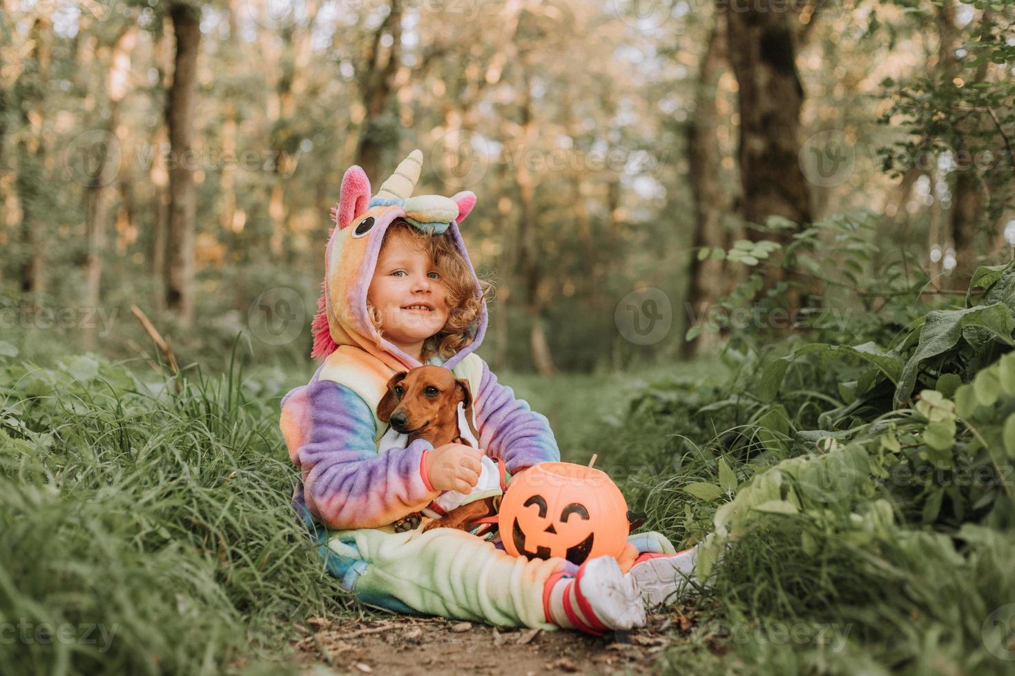 little girl in a rainbow unicorn Halloween costume and a dachshund in a dress with a pumpkin basket for sweets are sitting on stump at forest sunset. fabulous wonderful magical forest. space for text photo