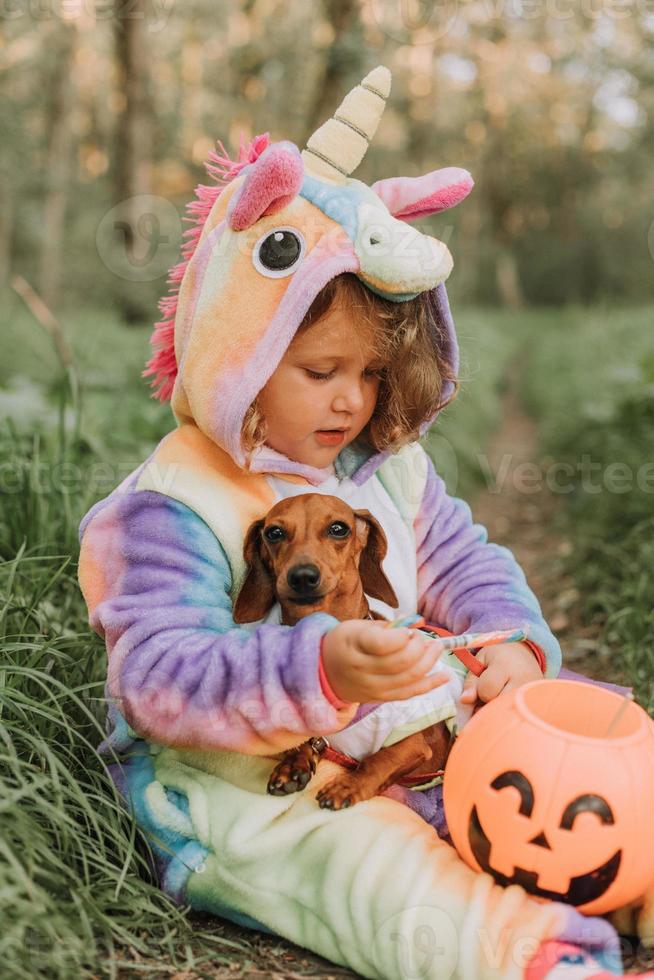 una niña con un disfraz de halloween de unicornio arcoíris y un dachshund con un vestido con una canasta de calabaza para dulces están sentados en un tocón al atardecer del bosque. fabuloso maravilloso bosque mágico. espacio para texto foto