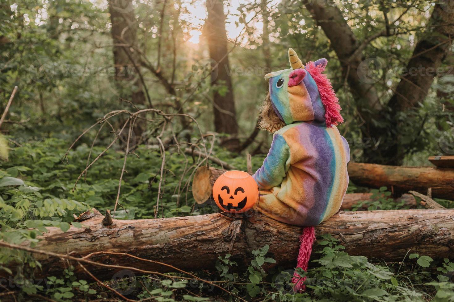 little girl in a rainbow unicorn Halloween costume with a pumpkin basket for sweets is sitting on a stump at a forest sunset. a fabulous wonderful magical forest. space for text. High quality photo