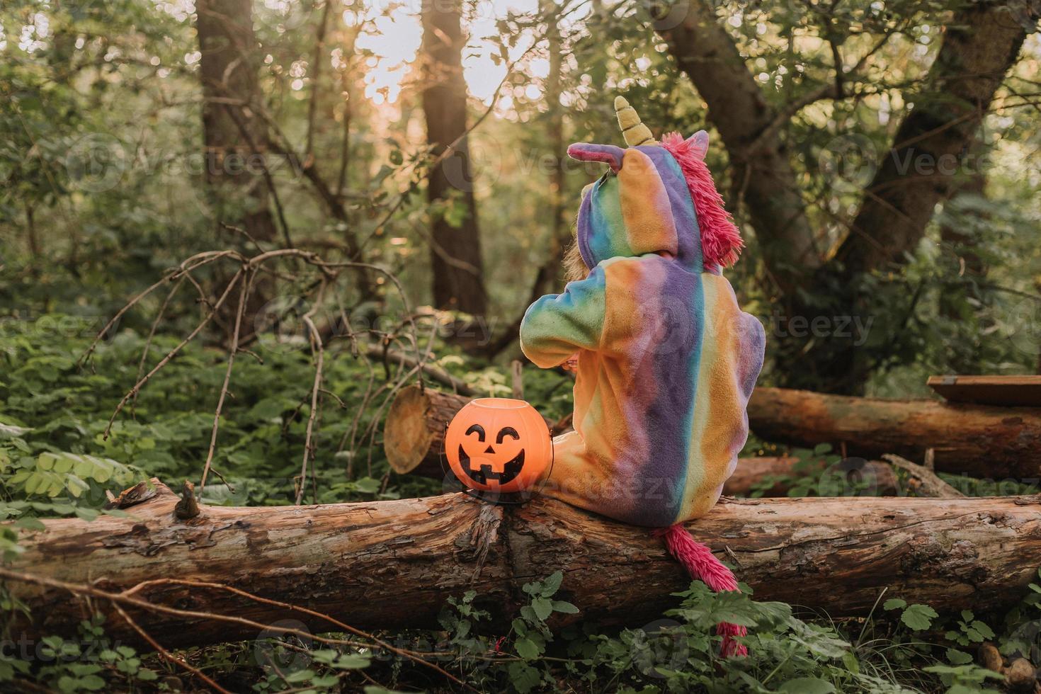 little girl in a rainbow unicorn Halloween costume with a pumpkin basket for sweets is sitting on a stump at a forest sunset. a fabulous wonderful magical forest. space for text. High quality photo