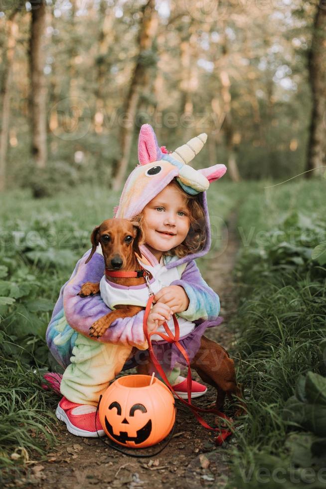 little girl in a rainbow unicorn Halloween costume and a dachshund in a dress with a pumpkin basket for sweets are sitting on stump at forest sunset. fabulous wonderful magical forest. space for text photo